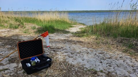 Instrument för vattenprover på en strand med vass, gräs, sand och vatten i bakgrunden